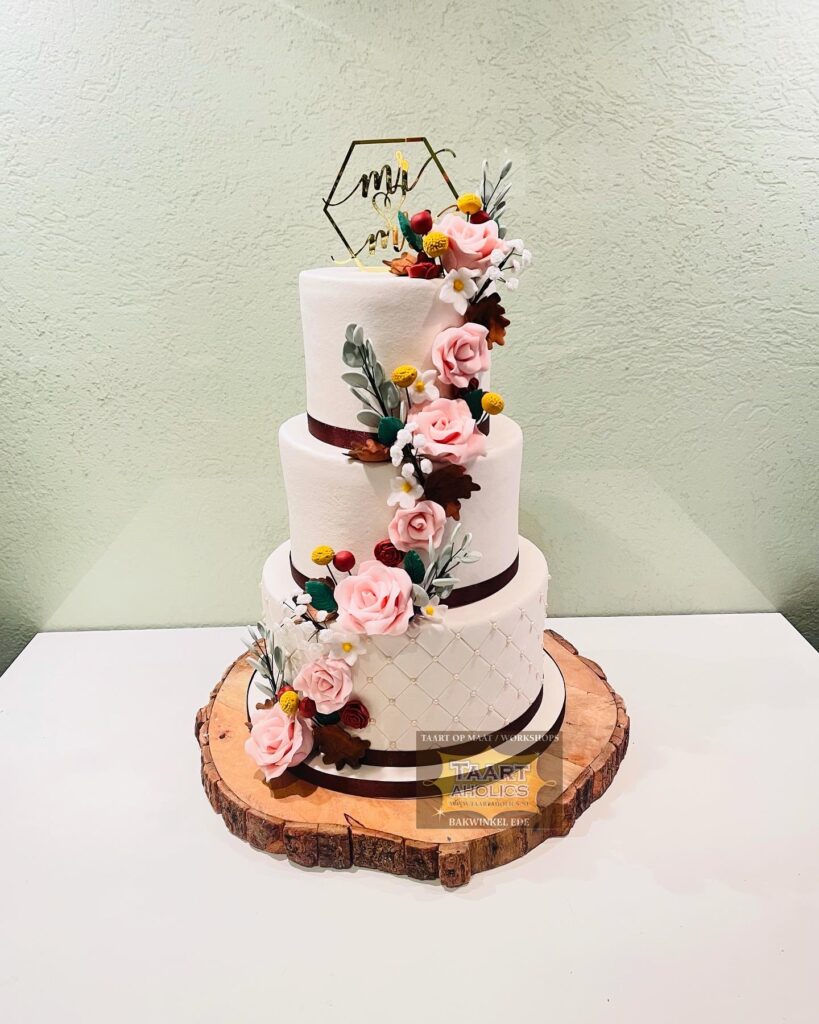 Drielaagse witte bruidstaart met suikerbloemen in roze en herfstkleuren, op een boomschijf